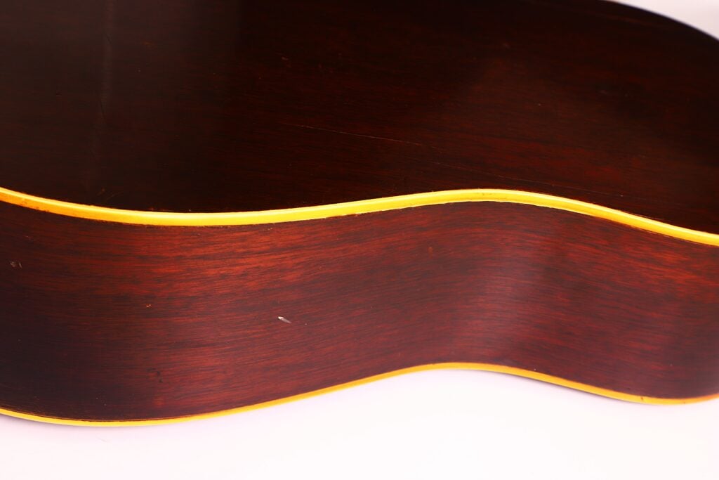 Nahaufnahme der Seite einer Gibson J-45 1954 Akustische Gitarre, die den dunkelbraunen Holzkorpus mit gelber Zierleiste, die sanfte Wölbung und die detaillierte Holzmaserung vor einem weißen Hintergrund zeigt. Nahaufnahme der Seite einer Gibson J-45 1954 Akustische Gitarre, die den dunkelbraunen Holzkorpus mit gelber Zierleiste, die sanfte Wölbung und die detaillierte Holzmaserung vor einem weißen Hintergrund zeigt.