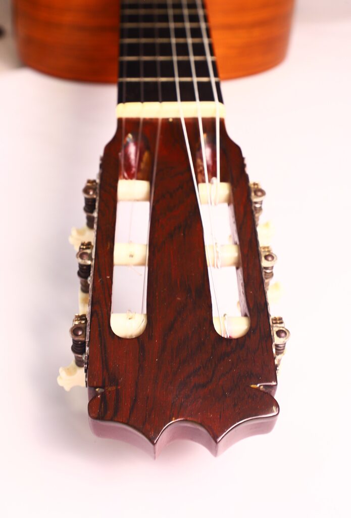Nahaufnahme der Kopfplatte einer Faustino Conde Negra Flamenco-Gitarre von Hermanos Conde aus dem Jahr 1971 mit Nylonsaiten, Stimmwirbeln und einer Holzoberfläche vor einem schlichten, hellen Hintergrund. Nahaufnahme der Kopfplatte einer Faustino Conde Negra Flamenco-Gitarre von Hermanos Conde aus dem Jahr 1971 mit Nylonsaiten, Stimmwirbeln und einer Holzoberfläche vor einem schlichten, hellen Hintergrund.