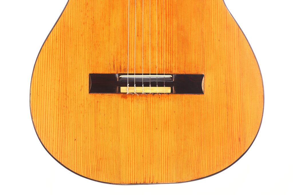 Nahaufnahme des Korpus der Juan Lopez Jimenez 1933 Klassische Gitarre. Hervorzuheben sind die Saiten, der Holzsteg und die natürliche Holzmaserung des Resonanzbodens vor einem weißen Hintergrund. Nahaufnahme des Korpus der Juan Lopez Jimenez 1933 Klassische Gitarre. Hervorzuheben sind die Saiten, der Holzsteg und die natürliche Holzmaserung des Resonanzbodens vor einem weißen Hintergrund.