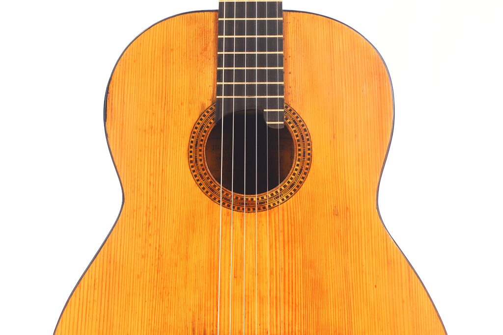Nahaufnahme von Korpus und Hals der Juan Lopez Jimenez 1933 Klassische Gitarre. Hervorzuheben sind die hölzerne Struktur, das runde Schallloch und die Nylonsaiten vor weißem Hintergrund. Nahaufnahme von Korpus und Hals der Juan Lopez Jimenez 1933 Klassische Gitarre. Hervorzuheben sind die hölzerne Struktur, das runde Schallloch und die Nylonsaiten vor weißem Hintergrund.