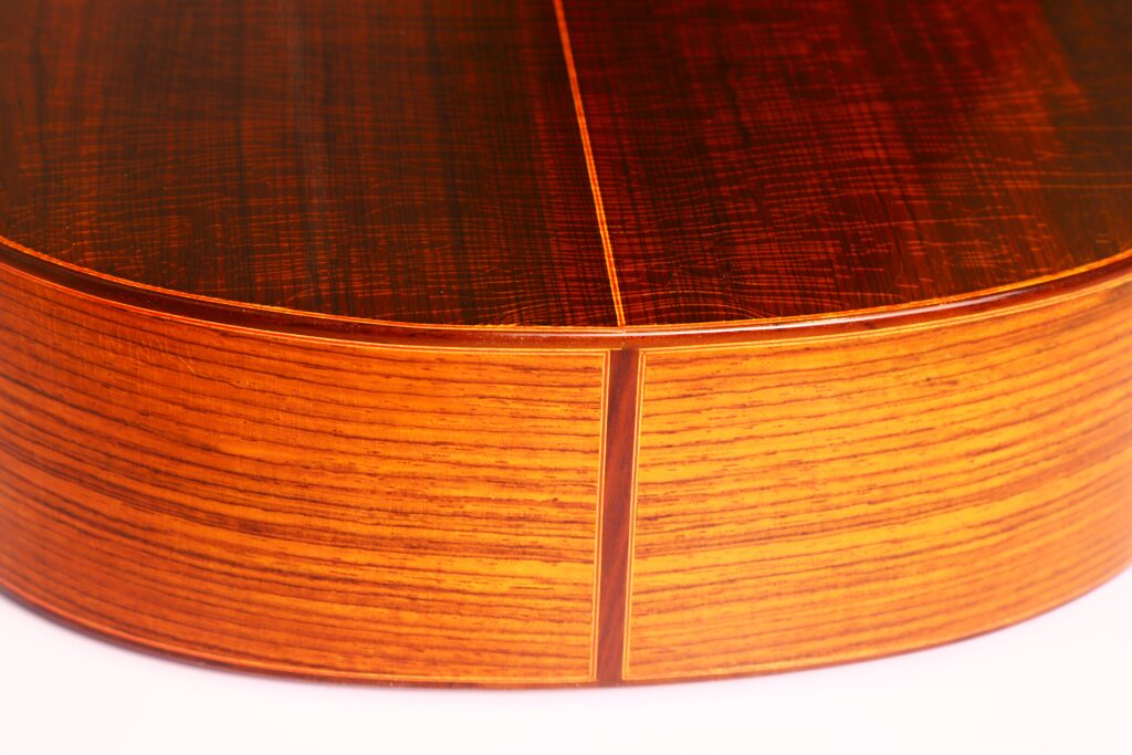 Nahaufnahme des Bodens und der Seite einer Faustino Conde Negra Flamenco-Gitarre von Hermanos Conde 1971. Hervorzuheben sind die natürliche Maserung des Holzes, eine Naht, an der sich zwei Teile treffen, und die warme orange-braune Lackierung. Nahaufnahme des Bodens und der Seite einer Faustino Conde Negra Flamenco-Gitarre von Hermanos Conde 1971. Hervorzuheben sind die natürliche Maserung des Holzes, eine Naht, an der sich zwei Teile treffen, und die warme orange-braune Lackierung.