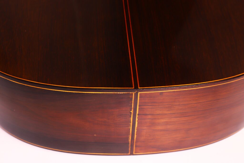 Nahaufnahme des Bodens und der Zargen einer Jose Ramirez 1a 1959 PB Klassische Gitarre. Das Holz ist dunkel poliert und hat eine glänzende Oberfläche mit feinen gelben Einlegearbeiten entlang der Nähte. Nahaufnahme des Bodens und der Zargen einer Jose Ramirez 1a 1959 PB Klassische Gitarre. Das Holz ist dunkel poliert und hat eine glänzende Oberfläche mit feinen gelben Einlegearbeiten entlang der Nähte.