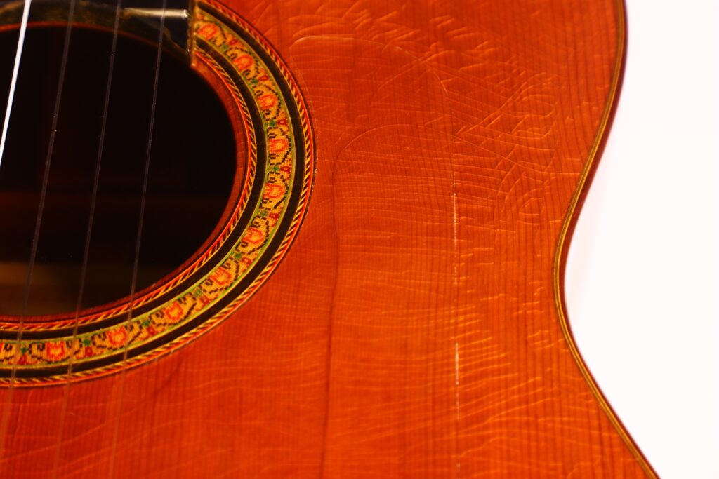 Nahaufnahme des glänzenden Korpus der Hermanos Conde 1971 Faustino Conde Negra Flamenco-Gitarre. Hervorzuheben sind die dekorative Rosette, die komplizierte Holzmaserung, das Schallloch und die teilweise sichtbaren Saiten auf der linken Seite. Nahaufnahme des glänzenden Korpus der Hermanos Conde 1971 Faustino Conde Negra Flamenco-Gitarre. Hervorzuheben sind die dekorative Rosette, die komplizierte Holzmaserung, das Schallloch und die teilweise sichtbaren Saiten auf der linken Seite.