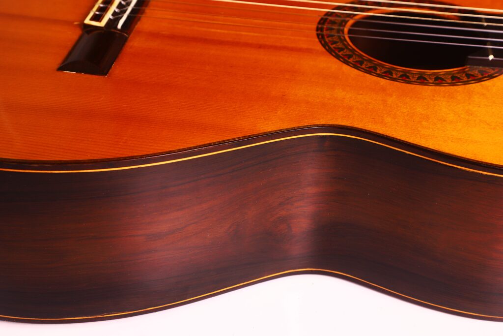 Nahaufnahme der Seite und der Decke einer Jose Ramirez 1a 1959 PB Klassische Gitarre. Hervorzuheben sind die Holzmaserung, die Saiten, das Schallloch mit Rosette und die glänzende Oberfläche. Nahaufnahme der Seite und der Decke einer Jose Ramirez 1a 1959 PB Klassische Gitarre. Hervorzuheben sind die Holzmaserung, die Saiten, das Schallloch mit Rosette und die glänzende Oberfläche.