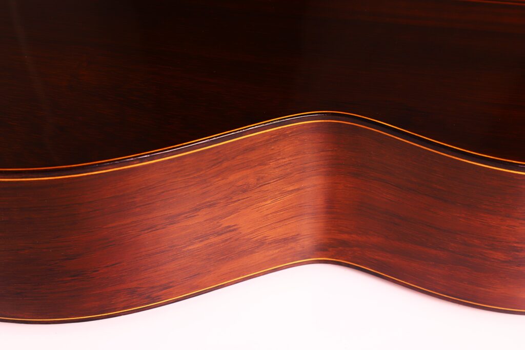 Nahaufnahme des Holzkorpus der Jose Ramirez 1a 1959 PB Klassische Gitarre. Hervorzuheben sind die glatte gewölbte Seite, die reiche braune Maserung und eine dünne kontrastierende Einlage entlang der Kante vor einem hellen Hintergrund. Nahaufnahme des Holzkorpus der Jose Ramirez 1a 1959 PB Klassische Gitarre. Hervorzuheben sind die glatte gewölbte Seite, die reiche braune Maserung und eine dünne kontrastierende Einlage entlang der Kante vor einem hellen Hintergrund.
