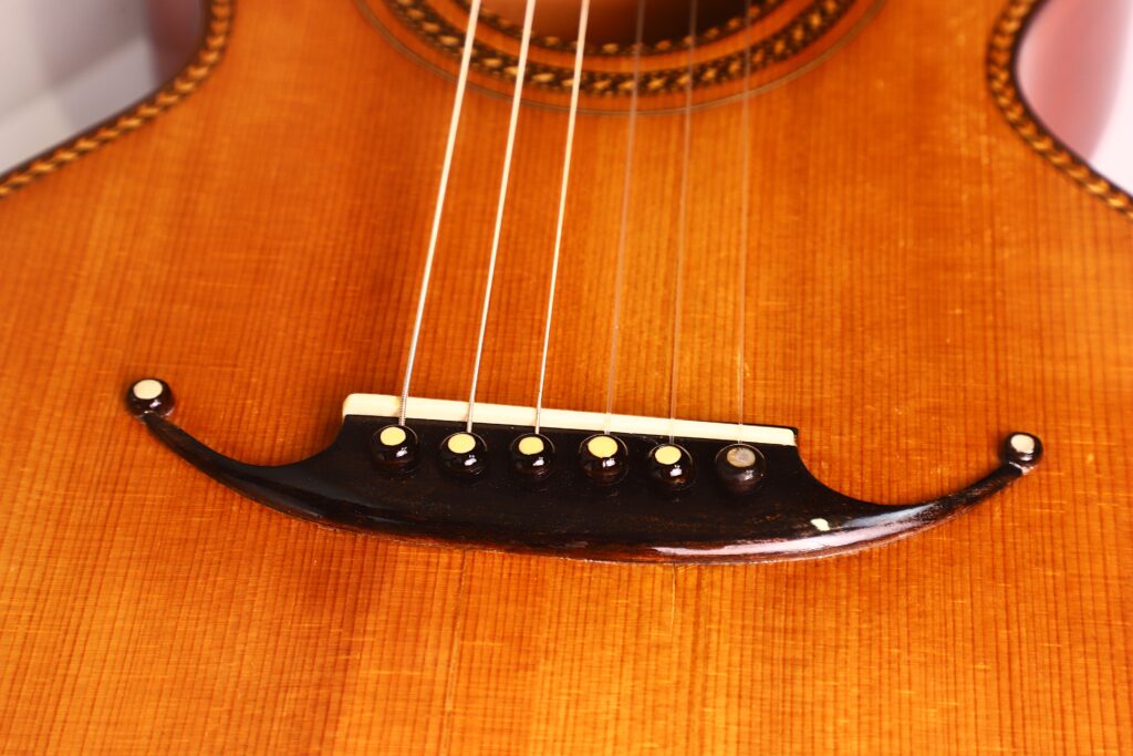 Nahaufnahme des Stegs und der Saiten der Marc Antonio Romantikgitarre. Sie zeigt dekorative Holzbögen mit sechs Saiten, die mit weißen Stiften auf einer glänzenden, sattbraunen Oberfläche verankert sind. Nahaufnahme des Stegs und der Saiten der Marc Antonio Romantikgitarre. Sie zeigt dekorative Holzbögen mit sechs Saiten, die mit weißen Stiften auf einer glänzenden, sattbraunen Oberfläche verankert sind.