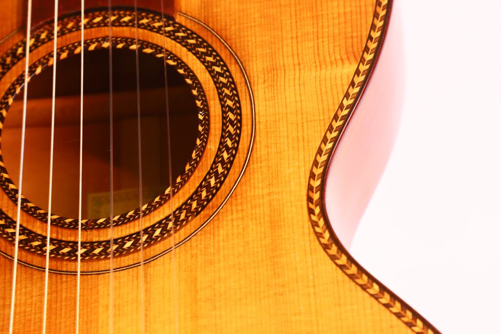 Nahaufnahme des Korpus der Marc Antonio Romantikgitarre mit detaillierter Holzmaserung, einer verzierten Rosette um das Schallloch und mehreren Gitarrensaiten vor einem hellen Hintergrund. Nahaufnahme des Korpus der Marc Antonio Romantikgitarre mit detaillierter Holzmaserung, einer verzierten Rosette um das Schallloch und mehreren Gitarrensaiten vor einem hellen Hintergrund.