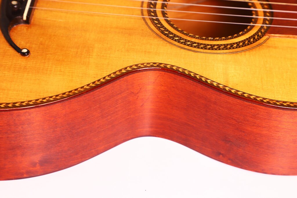 Nahaufnahme des hölzernen Korpus der Marc Antonio Romantikgitarre, wobei die detaillierte Maserung, die verzierte Rosette und die dekorative Randeinlage vor einem weißen Hintergrund hervorgehoben werden. Nahaufnahme des hölzernen Korpus der Marc Antonio Romantikgitarre, wobei die detaillierte Maserung, die verzierte Rosette und die dekorative Randeinlage vor einem weißen Hintergrund hervorgehoben werden.