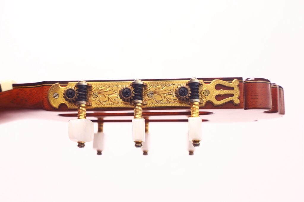 Seitliche Nahaufnahme der Kopfplatte der Manuel Contreras Double Top 1985 Klassische Gitarre, mit verschnörkelten goldenen Mechaniken mit schwarzen und weißen Knöpfen vor einem schlichten weißen Hintergrund. Seitliche Nahaufnahme der Kopfplatte der Manuel Contreras Double Top 1985 Klassische Gitarre, mit verschnörkelten goldenen Mechaniken mit schwarzen und weißen Knöpfen vor einem schlichten weißen Hintergrund.