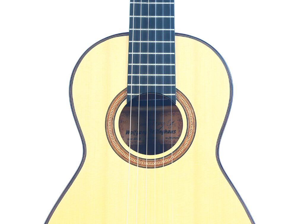 Nahaufnahme der Jellinghaus Torres 77 PU Klassische Gitarre. Hervorzuheben sind der helle Holzkorpus und -hals, die Metallsaiten und die verzierte Rosette um das Schallloch. Nahaufnahme der Jellinghaus Torres 77 PU Klassische Gitarre. Hervorzuheben sind der helle Holzkorpus und -hals, die Metallsaiten und die verzierte Rosette um das Schallloch.