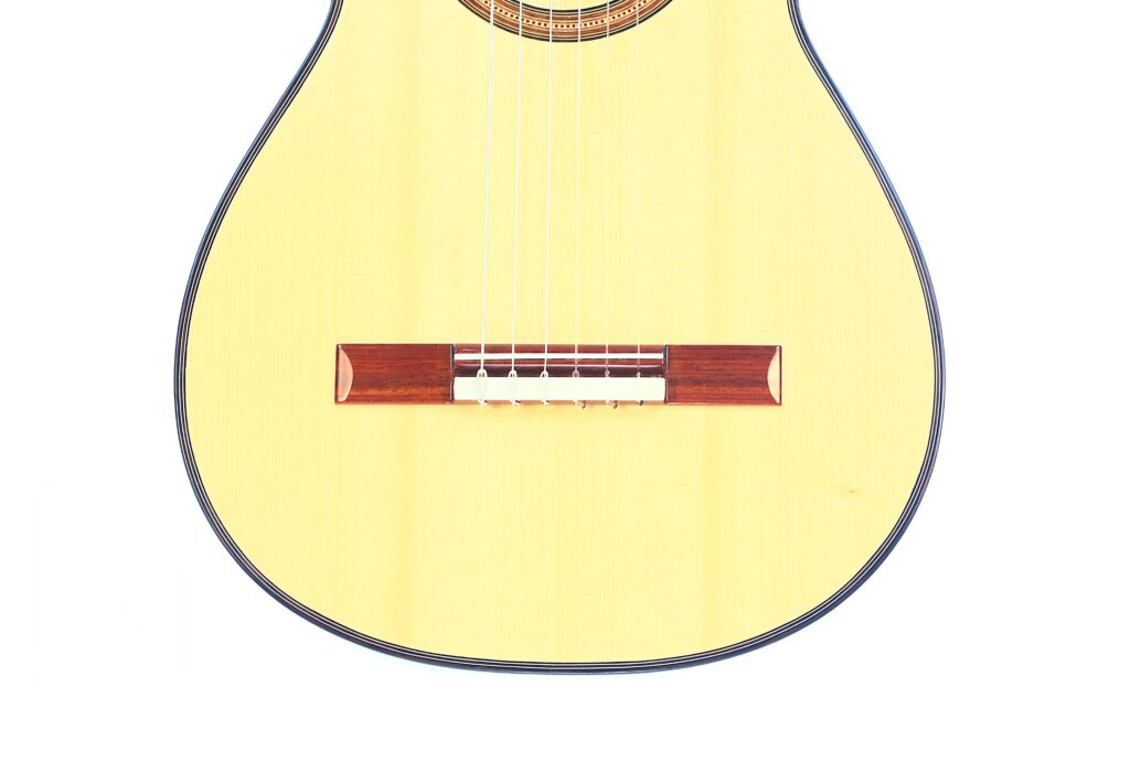 Nahaufnahme der unteren Hälfte der Jellinghaus Torres 77 PU Klassische Gitarre. Hervorzuheben sind der helle Holzkorpus, die Saiten und der dunkle Holzsteg vor einem weißen Hintergrund. Nahaufnahme der unteren Hälfte der Jellinghaus Torres 77 PU Klassische Gitarre. Hervorzuheben sind der helle Holzkorpus, die Saiten und der dunkle Holzsteg vor einem weißen Hintergrund.
