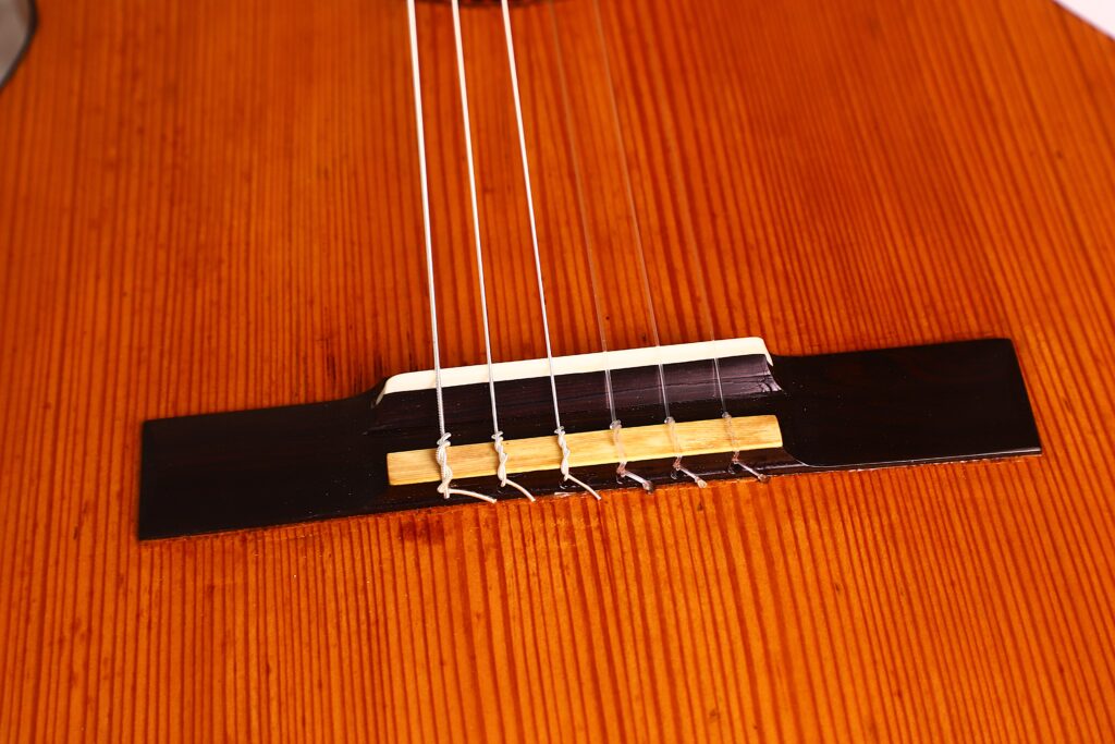 Nahaufnahme des Stegs der Juan Lopez Jimenez 1933 Klassische Gitarre mit sechs Saiten auf einem polierten braunen Holzkorpus mit sichtbaren Holzmaserungen. Nahaufnahme des Stegs der Juan Lopez Jimenez 1933 Klassische Gitarre mit sechs Saiten auf einem polierten braunen Holzkorpus mit sichtbaren Holzmaserungen.