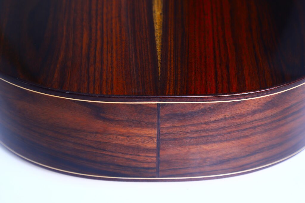 Nahaufnahme der Rückseite und der Seite der Jellinghaus Torres 77 PU Klassische Gitarre, die die glatte, polierte Holzmaserung mit glänzender Oberfläche und feinen Einlegearbeiten zeigt. Nahaufnahme der Rückseite und der Seite der Jellinghaus Torres 77 PU Klassische Gitarre, die die glatte, polierte Holzmaserung mit glänzender Oberfläche und feinen Einlegearbeiten zeigt.