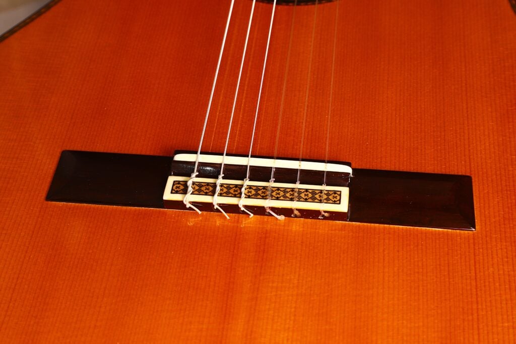 Nahaufnahme des Stegs und der Saiten der Manuel Contreras Double Top 1985 Klassische Gitarre, die die detaillierten Einlegearbeiten und die glänzende Holzoberfläche zeigt. Nahaufnahme des Stegs und der Saiten der Manuel Contreras Double Top 1985 Klassische Gitarre, die die detaillierten Einlegearbeiten und die glänzende Holzoberfläche zeigt.