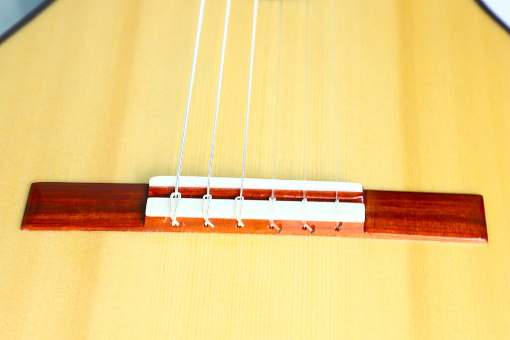 Nahaufnahme von Steg und Saiten der Jellinghaus Torres 77 PU Klassische Gitarre mit Holzsteg, weißem Sattel und Saitenknoten auf einem hellen Korpus. Nahaufnahme von Steg und Saiten der Jellinghaus Torres 77 PU Klassische Gitarre mit Holzsteg, weißem Sattel und Saitenknoten auf einem hellen Korpus.