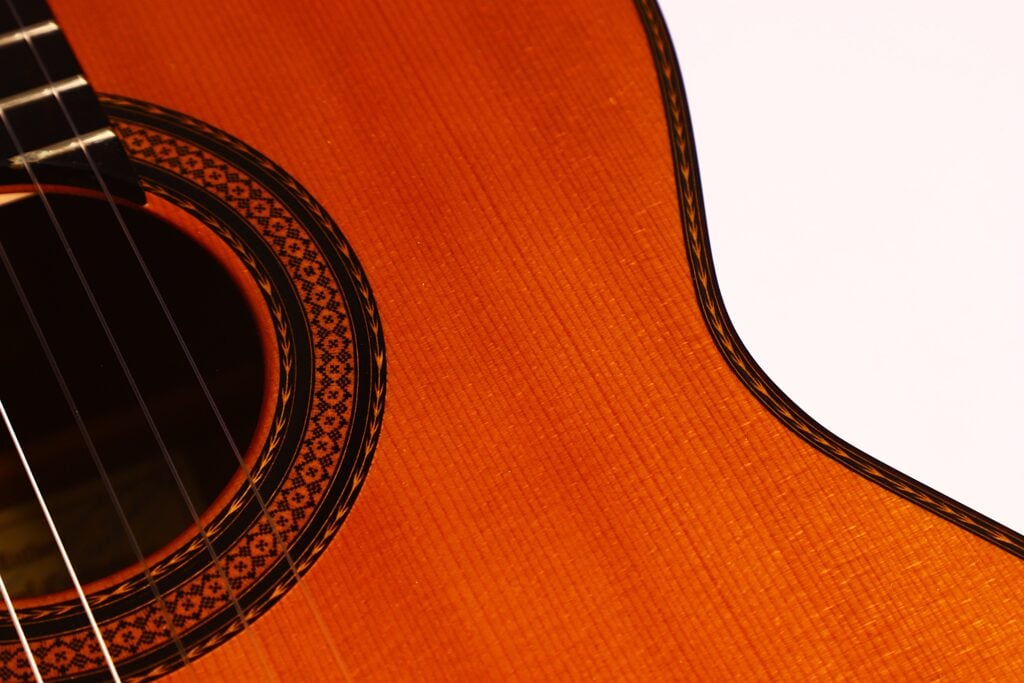 Nahaufnahme einer Manuel Contreras Double Top 1985 Klassische Gitarre, die das Schallloch mit dekorativer Rosette, die Saiten und den polierten Holzkorpus vor einem weißen Hintergrund zeigt. Nahaufnahme einer Manuel Contreras Double Top 1985 Klassische Gitarre, die das Schallloch mit dekorativer Rosette, die Saiten und den polierten Holzkorpus vor einem weißen Hintergrund zeigt.
