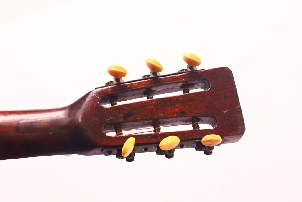 Nahaufnahme der Kopfplatte einer Martin 00-21 Akustikgitarre von 1931 mit sechs Stimmwirbeln mit gelben Knöpfen auf dunklem Holz vor einem schlichten weißen Hintergrund. Nahaufnahme der Kopfplatte einer Martin 00-21 Akustikgitarre von 1931 mit sechs Stimmwirbeln mit gelben Knöpfen auf dunklem Holz vor einem schlichten weißen Hintergrund.