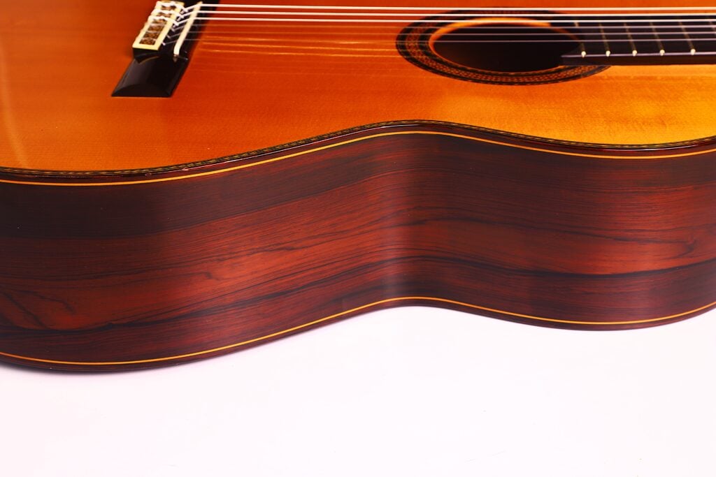 Eine Nahaufnahme der Seite und des unteren Korpus der Manuel Contreras Double Top 1985 Klassische Gitarre zeigt die glänzende Holzoberfläche, die sichtbare Maserung und die eleganten Kurven vor einem weißen Hintergrund. Eine Nahaufnahme der Seite und des unteren Korpus der Manuel Contreras Double Top 1985 Klassische Gitarre zeigt die glänzende Holzoberfläche, die sichtbare Maserung und die eleganten Kurven vor einem weißen Hintergrund.