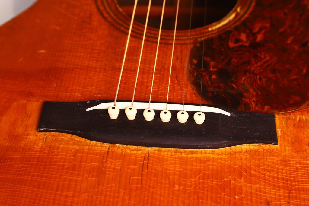 Nahaufnahme des Stegs der Martin 00-21 1931 Akustik-Gitarre mit sechs Saiten, die an elfenbeinfarbenen Stiften auf einem schwarzen Holzsteg befestigt sind, wobei ein Teil des rotbraunen Korpus und des Schlagbretts zu sehen ist. Nahaufnahme des Stegs der Martin 00-21 1931 Akustik-Gitarre mit sechs Saiten, die an elfenbeinfarbenen Stiften auf einem schwarzen Holzsteg befestigt sind, wobei ein Teil des rotbraunen Korpus und des Schlagbretts zu sehen ist.
