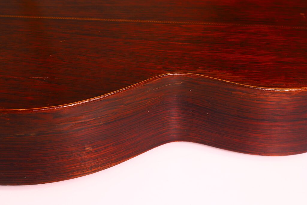 Nahaufnahme der Seite und der Rückseite einer Martin 00-21 1931 Akustik Gitarre, die das glatte, polierte, rötlich-braune Holz und die eleganten Kurven vor einem hellen Hintergrund hervorhebt. Nahaufnahme der Seite und der Rückseite einer Martin 00-21 1931 Akustik Gitarre, die das glatte, polierte, rötlich-braune Holz und die eleganten Kurven vor einem hellen Hintergrund hervorhebt.