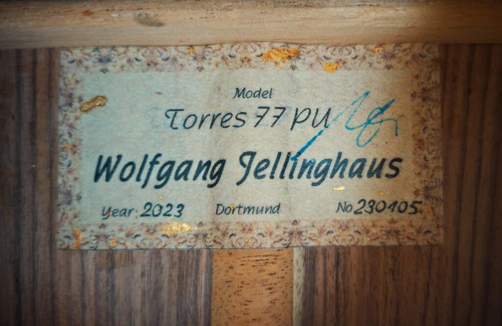 Eine Nahaufnahme des Gitarrenetiketts zeigt: Jellinghaus Torres 77 PU Klassische Gitarre, Wolfgang Jellinghaus, Jahr: 2023, Dortmund, Nr: 230405, mit handschriftlichen blauen Tintenmarkierungen auf einer Holzoberfläche. Eine Nahaufnahme des Gitarrenetiketts zeigt: Jellinghaus Torres 77 PU Klassische Gitarre, Wolfgang Jellinghaus, Jahr: 2023, Dortmund, Nr: 230405, mit handschriftlichen blauen Tintenmarkierungen auf einer Holzoberfläche.