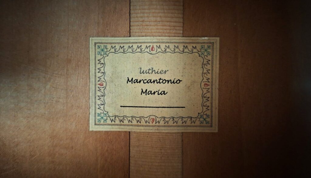 Nahaufnahme eines Etiketts im Inneren der Marc Antonio Romantikgitarre mit der Aufschrift "luthier Marcantonio María" in schwarzer Schrift und einem dekorativen Rahmen mit kleinen roten und grünen Details. Nahaufnahme eines Etiketts im Inneren der Marc Antonio Romantikgitarre mit der Aufschrift "luthier Marcantonio María" in schwarzer Schrift und einem dekorativen Rahmen mit kleinen roten und grünen Details.