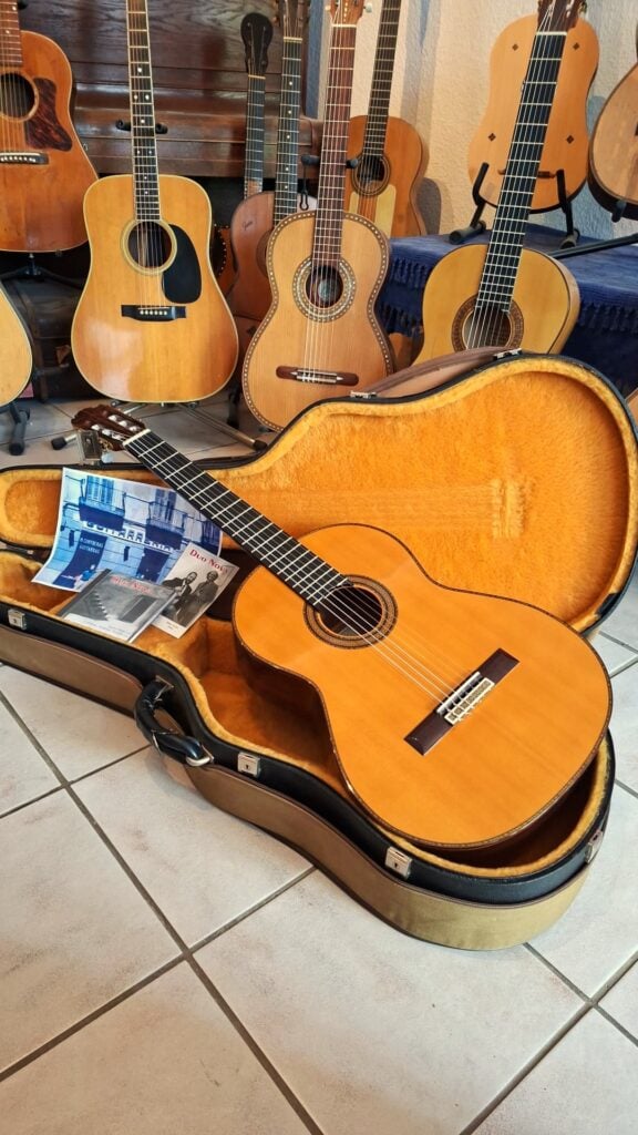 Eine Manuel Contreras Double Top 1985 Klassische Gitarre liegt in einem offenen, mit Plüsch gefütterten Hartschalenkoffer auf einem gefliesten Boden, umgeben von anderen Akustikgitarren auf Ständern, mit einem Blatt Papier oder einem Heft im Inneren des Koffers. Eine Manuel Contreras Double Top 1985 Klassische Gitarre liegt in einem offenen, mit Plüsch gefütterten Hartschalenkoffer auf einem gefliesten Boden, umgeben von anderen Akustikgitarren auf Ständern, mit einem Blatt Papier oder einem Heft im Inneren des Koffers.