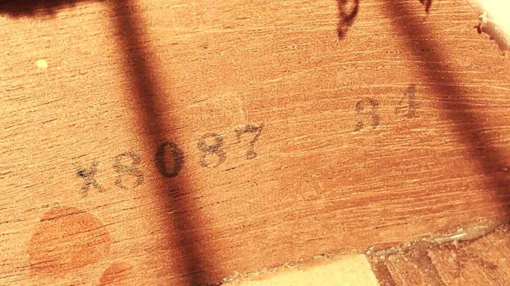 Nahaufnahme der Holzoberfläche einer Gibson J-45 1954 Akustische Gitarre, auf der die Nummern 18087 84 in verblasster schwarzer Tinte eingeprägt sind, zusammen mit sichtbaren Schatten und Spuren auf dem Holz. Nahaufnahme der Holzoberfläche einer Gibson J-45 1954 Akustische Gitarre, auf der die Nummern 18087 84 in verblasster schwarzer Tinte eingeprägt sind, zusammen mit sichtbaren Schatten und Spuren auf dem Holz.