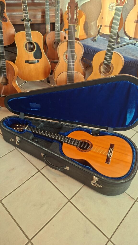 Eine Jose Ramirez 1a 1959 PB Klassische Gitarre liegt in einem offenen, blau ausgekleideten Hartschalenkoffer auf einem gefliesten Boden, während im Hintergrund weitere akustische und klassische Gitarren aufrecht stehen. Eine Jose Ramirez 1a 1959 PB Klassische Gitarre liegt in einem offenen, blau ausgekleideten Hartschalenkoffer auf einem gefliesten Boden, während im Hintergrund weitere akustische und klassische Gitarren aufrecht stehen.