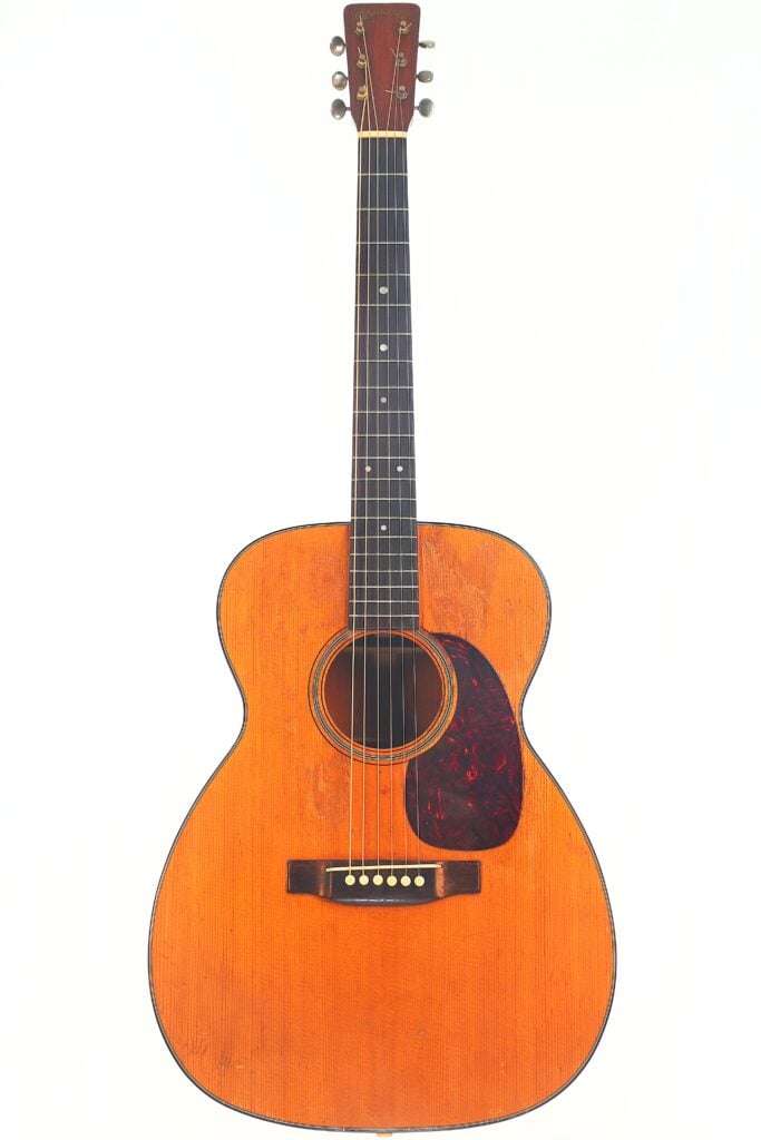 Die Martin 00-18 Akustik Gitarre aus dem Jahr 1954 verfügt über eine naturbelassene Holzoberfläche, ein schwarzes Schlagbrett, sechs Saiten und Punkt-Einlagen auf dem Griffbrett; sie ist aufrecht vor einem schlichten weißen Hintergrund abgebildet. Die Martin 00-18 Akustik Gitarre aus dem Jahr 1954 verfügt über eine naturbelassene Holzoberfläche, ein schwarzes Schlagbrett, sechs Saiten und Punkt-Einlagen auf dem Griffbrett; sie ist aufrecht vor einem schlichten weißen Hintergrund abgebildet.
