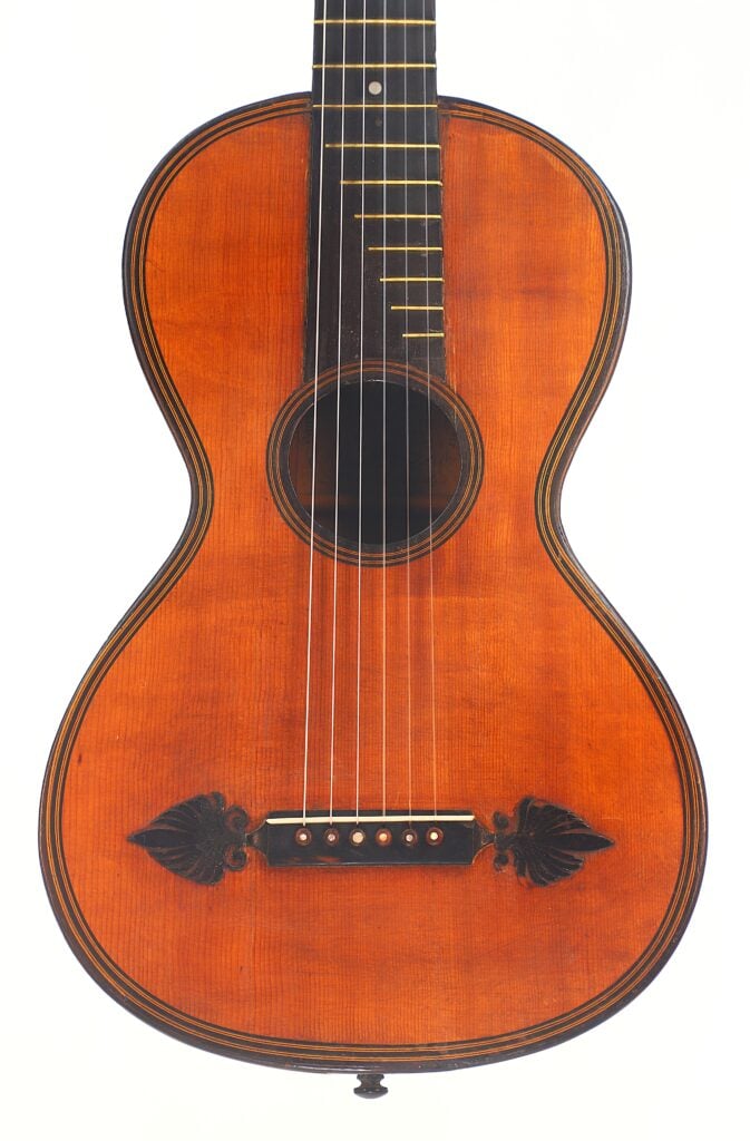 Eine Nahaufnahme der Romantikgitarre von Stephan Thumhart aus dem Jahr 1838, die sich durch einen rotbraunen Holzkorpus, dunkle Zierdetails um das Schallloch und den Steg sowie blattförmige Verzierungen in der Nähe des Stegs auszeichnet. Eine Nahaufnahme der Romantikgitarre von Stephan Thumhart aus dem Jahr 1838, die sich durch einen rotbraunen Holzkorpus, dunkle Zierdetails um das Schallloch und den Steg sowie blattförmige Verzierungen in der Nähe des Stegs auszeichnet.