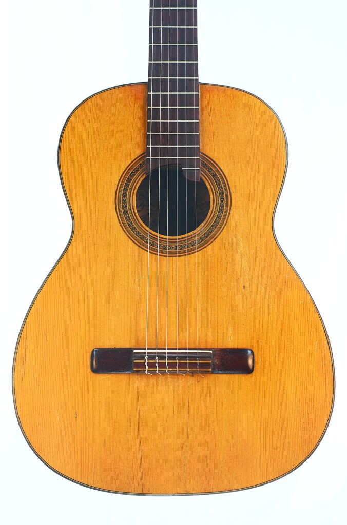 Die klassische Gitarre „Jose Ramirez I 1921“ verfügt über eine naturbelassene Holzoberfläche, sechs Saiten, ein rundes Schallloch mit dekorativer Rosette sowie einen dunklen Holzsteg und wird vor einem schlichten weißen Hintergrund präsentiert. Die klassische Gitarre „Jose Ramirez I 1921“ verfügt über eine naturbelassene Holzoberfläche, sechs Saiten, ein rundes Schallloch mit dekorativer Rosette sowie einen dunklen Holzsteg und wird vor einem schlichten weißen Hintergrund präsentiert.