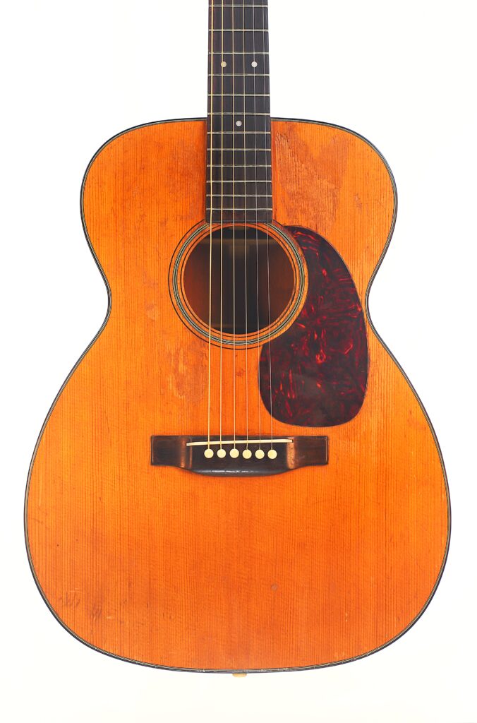 Eine Nahaufnahme der Martin 00-18 Akustikgitarre aus dem Jahr 1954 mit naturbelassener Holzoberfläche, dunklem Schlagbrett und sichtbaren Saiten, fotografiert vor weißem Hintergrund. Eine Nahaufnahme der Martin 00-18 Akustikgitarre aus dem Jahr 1954 mit naturbelassener Holzoberfläche, dunklem Schlagbrett und sichtbaren Saiten, fotografiert vor weißem Hintergrund.