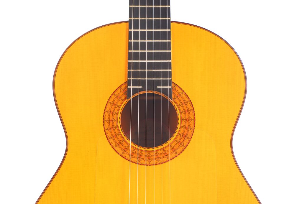 Nahaufnahme des oberen Korpus und des Schalllochs einer Juan Montero 1993 Blanca Flamenco-Gitarre, mit Darstellung der Saiten, des Griffbretts und der verzierten Rosette um das Schallloch, vor einem weißen Hintergrund. Nahaufnahme des oberen Korpus und des Schalllochs einer Juan Montero 1993 Blanca Flamenco-Gitarre, mit Darstellung der Saiten, des Griffbretts und der verzierten Rosette um das Schallloch, vor einem weißen Hintergrund.