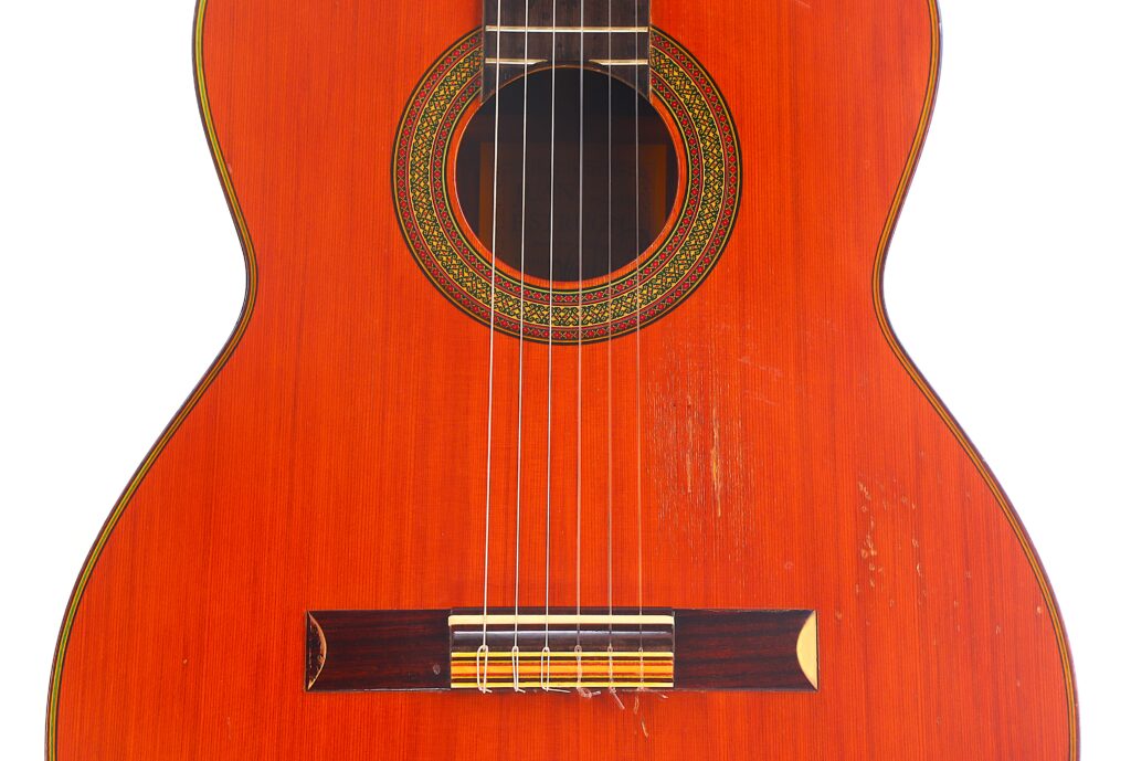 Nahaufnahme eines Korpus einer Juan Estruch Chet Atkins Yellow Label 1976 Klassische Gitarre mit rotbrauner Holzoberfläche, sechs Saiten, einem verzierten Schallloch und sichtbaren Kratzspuren auf der Oberfläche. Nahaufnahme eines Korpus einer Juan Estruch Chet Atkins Yellow Label 1976 Klassische Gitarre mit rotbrauner Holzoberfläche, sechs Saiten, einem verzierten Schallloch und sichtbaren Kratzspuren auf der Oberfläche.