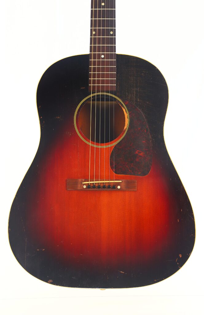 Nahaufnahme einer Gibson J-45 Banner 1944 Akustik-Gitarre mit Sunburst-Lackierung, die Gebrauchsspuren und Kratzer am Korpus aufweist. Das Bild konzentriert sich auf das Schallloch, Brücke und Griffbrett vor einem weißen Hintergrund. Nahaufnahme einer Gibson J-45 Banner 1944 Akustik-Gitarre mit Sunburst-Lackierung, die Gebrauchsspuren und Kratzer am Korpus aufweist. Das Bild konzentriert sich auf das Schallloch, Brücke und Griffbrett vor einem weißen Hintergrund.