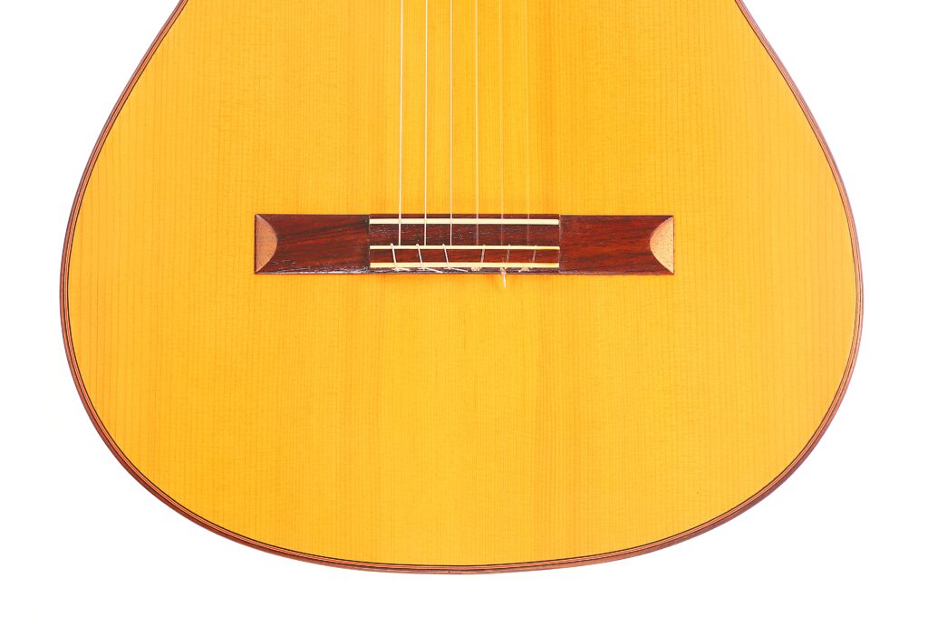 Nahaufnahme der unteren Hälfte einer „Carl Hermann Schäfer – Nicolaus Wollf 2012 Klassische Gitarre“, die den Holzkorpus, die sechs Saiten und den dunklen Steg vor weißem Hintergrund zeigt. Nahaufnahme der unteren Hälfte einer „Carl Hermann Schäfer – Nicolaus Wollf 2012 Klassische Gitarre“, die den Holzkorpus, die sechs Saiten und den dunklen Steg vor weißem Hintergrund zeigt.