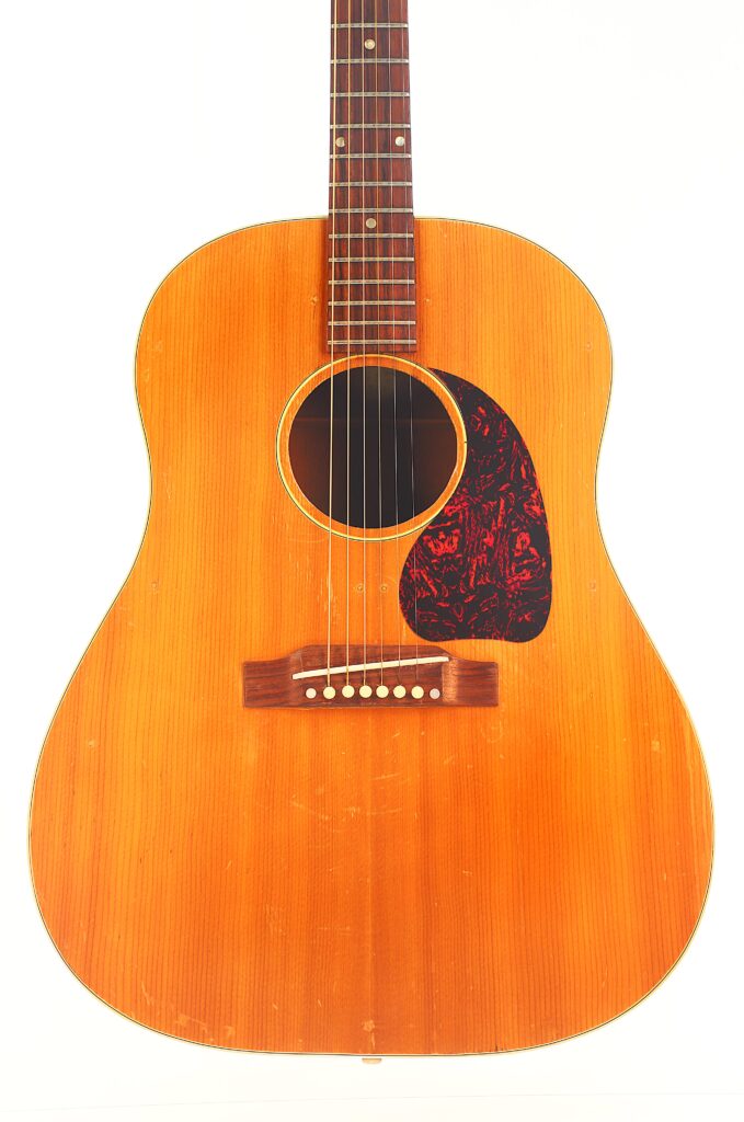 Eine Vorderansicht der Gibson J-50 Akustikgitarre aus dem Jahr 1952 zeigt ihre Naturholz-Oberfläche, das runde Schallloch, das dunkelrote, marmorierte Schlagbrett und die sechs Saiten. Eine Vorderansicht der Gibson J-50 Akustikgitarre aus dem Jahr 1952 zeigt ihre Naturholz-Oberfläche, das runde Schallloch, das dunkelrote, marmorierte Schlagbrett und die sechs Saiten.