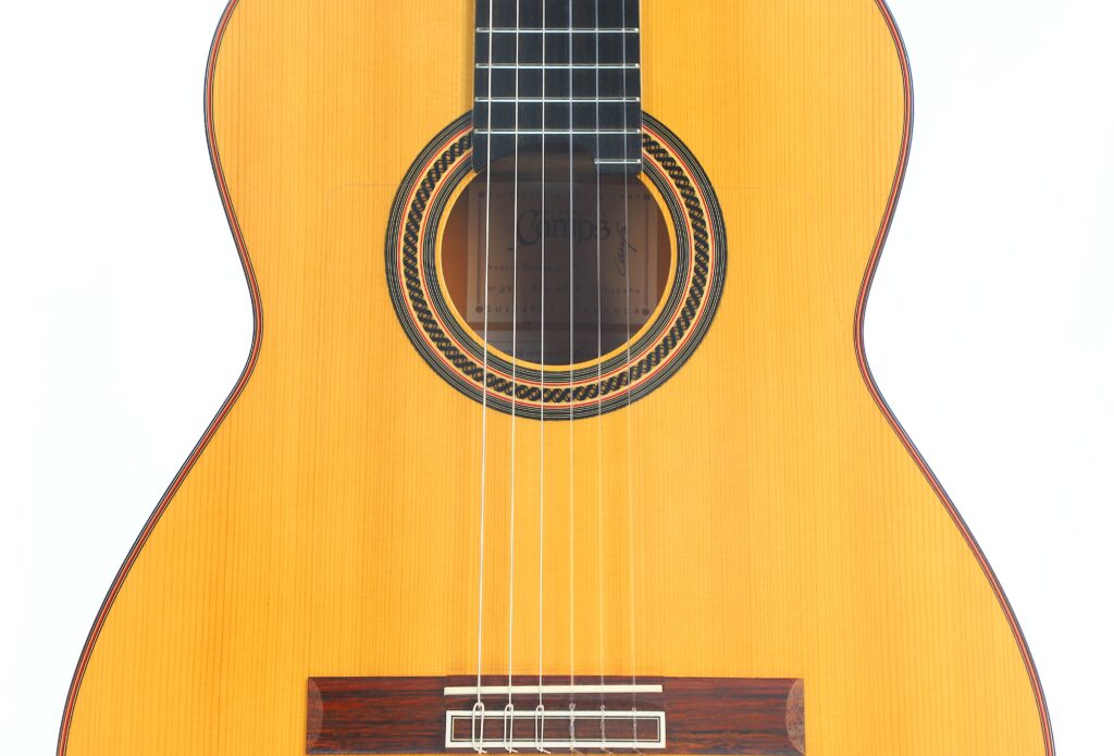 Nahaufnahme der Vorderseite der Flamenco-Gitarre „Camps Primera A 2018“, auf der die Saiten, das Schallloch mit der dekorativen Rosette sowie ein Teil des Holzkorpus vor weißem Hintergrund zu sehen sind. Nahaufnahme der Vorderseite der Flamenco-Gitarre „Camps Primera A 2018“, auf der die Saiten, das Schallloch mit der dekorativen Rosette sowie ein Teil des Holzkorpus vor weißem Hintergrund zu sehen sind.
