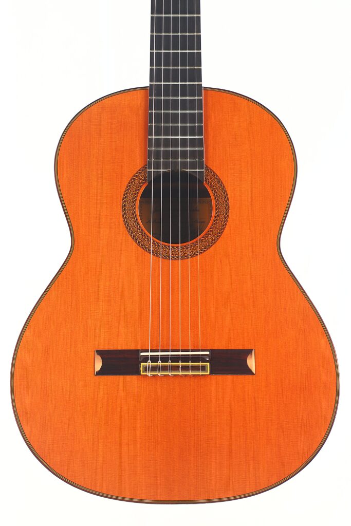 Die Juan Miguel Gonzalez 2006 Klassische Gitarre ist eine klassische Akustikgitarre mit einem rötlich-braunen Holzkorpus, sechs Saiten, einer dekorativen Rosette und einem dunklen Griffbrett, das vor einem schlichten weißen Hintergrund steht. Die Juan Miguel Gonzalez 2006 Klassische Gitarre ist eine klassische Akustikgitarre mit einem rötlich-braunen Holzkorpus, sechs Saiten, einer dekorativen Rosette und einem dunklen Griffbrett, das vor einem schlichten weißen Hintergrund steht.