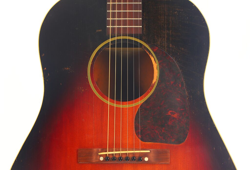 Nahaufnahme einer Gibson J-45 Banner 1944 Akustik Gitarre mit Sunburst-Lackierung, sechs Saiten, Schildpatt-Schlagbrett und Holzsteg mit weißen Stegstiften vor weißem Hintergrund. Nahaufnahme einer Gibson J-45 Banner 1944 Akustik Gitarre mit Sunburst-Lackierung, sechs Saiten, Schildpatt-Schlagbrett und Holzsteg mit weißen Stegstiften vor weißem Hintergrund.