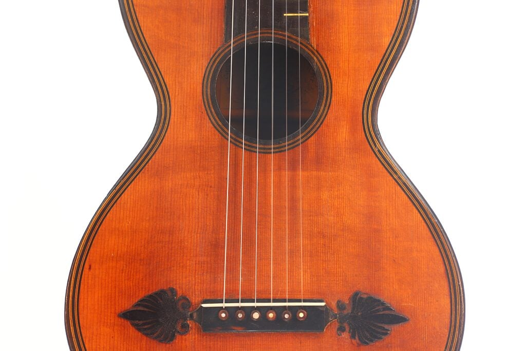 Nahaufnahme des Holzkörpers der „Romantikgitarre“ von Stephan Thumhart aus dem Jahr 1838, die die schwarzen Verzierungen im Bereich des Stegs und der Saiten vor einem weißen Hintergrund hervorhebt. Nahaufnahme des Holzkörpers der „Romantikgitarre“ von Stephan Thumhart aus dem Jahr 1838, die die schwarzen Verzierungen im Bereich des Stegs und der Saiten vor einem weißen Hintergrund hervorhebt.
