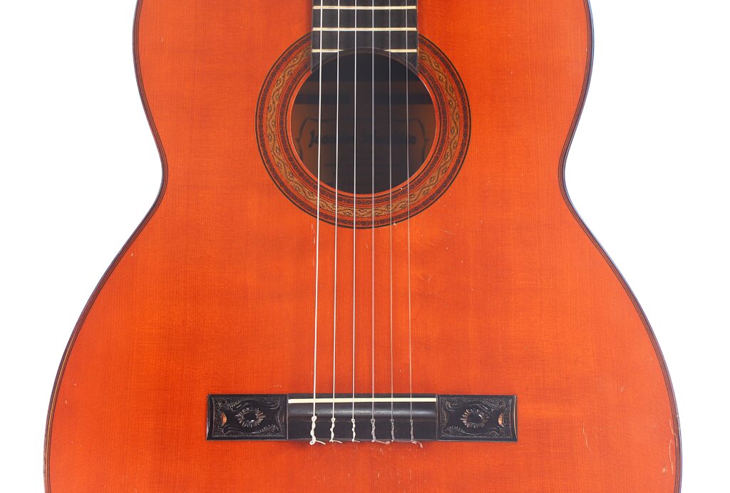 Nahaufnahme einer „Klassische Gitarre“ von Joachim Schneider aus dem Jahr 1977, auf der die Saiten, die dekorative Rosette um das Schallloch sowie ein Teil des kunstvoll verzierten Holzkörpers mit dem detailreichen Steg zu sehen sind. Nahaufnahme einer „Klassische Gitarre“ von Joachim Schneider aus dem Jahr 1977, auf der die Saiten, die dekorative Rosette um das Schallloch sowie ein Teil des kunstvoll verzierten Holzkörpers mit dem detailreichen Steg zu sehen sind.