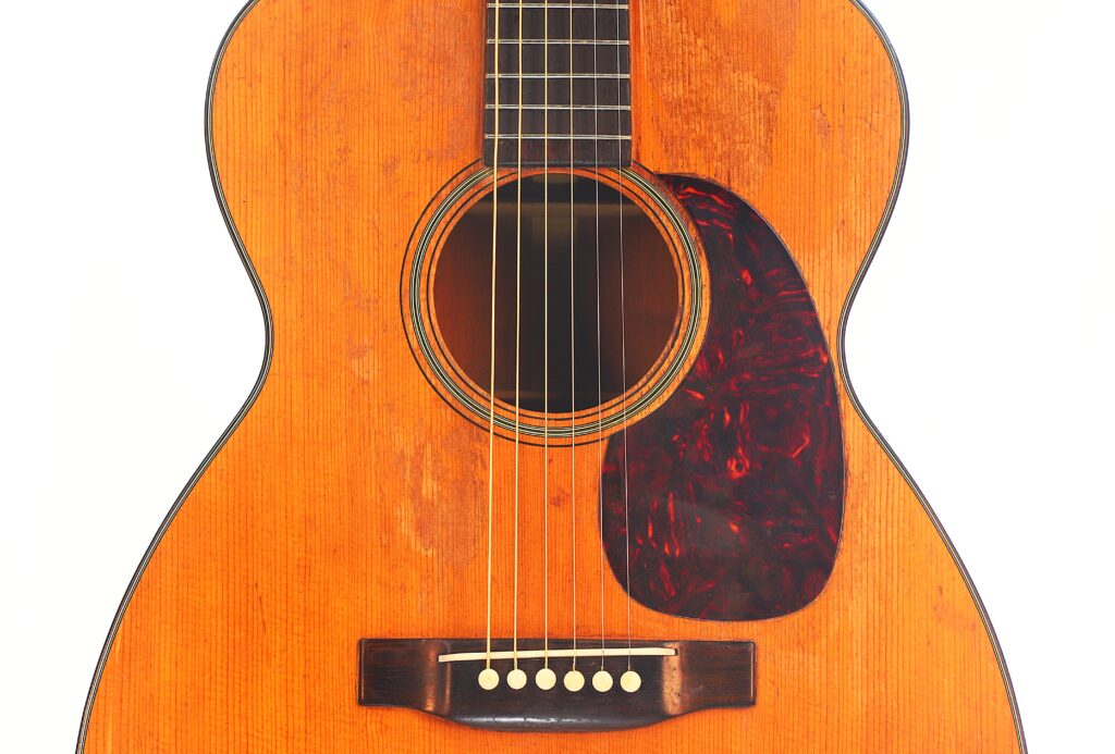 Nahaufnahme einer Martin 00-18 Akustikgitarre aus dem Jahr 1954, die ihre Holzoberfläche, die sechs Saiten, das dunkle Schlagbrett mit Schildpattmuster und den Steg mit weißen Stiften vor weißem Hintergrund zeigt. Nahaufnahme einer Martin 00-18 Akustikgitarre aus dem Jahr 1954, die ihre Holzoberfläche, die sechs Saiten, das dunkle Schlagbrett mit Schildpattmuster und den Steg mit weißen Stiften vor weißem Hintergrund zeigt.