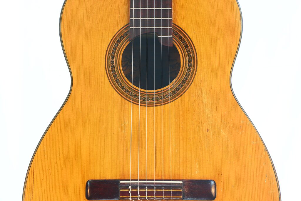Nahaufnahme der klassischen Gitarre „Jose Ramirez I 1921“, bei der der Korpus, die Saiten, das Schallloch, das Rosettenmuster und der Holzsteg vor weißem Hintergrund besonders hervorgehoben sind. Nahaufnahme der klassischen Gitarre „Jose Ramirez I 1921“, bei der der Korpus, die Saiten, das Schallloch, das Rosettenmuster und der Holzsteg vor weißem Hintergrund besonders hervorgehoben sind.