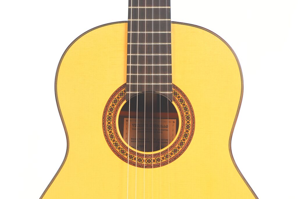 Eine Nahaufnahme des Korpus und der Saiten einer Vicente Carrillo 1998 Torres Klassische Gitarre, mit einer dekorativen Rosette um das Schallloch, fotografiert vor einem weißen Hintergrund. Eine Nahaufnahme des Korpus und der Saiten einer Vicente Carrillo 1998 Torres Klassische Gitarre, mit einer dekorativen Rosette um das Schallloch, fotografiert vor einem weißen Hintergrund.