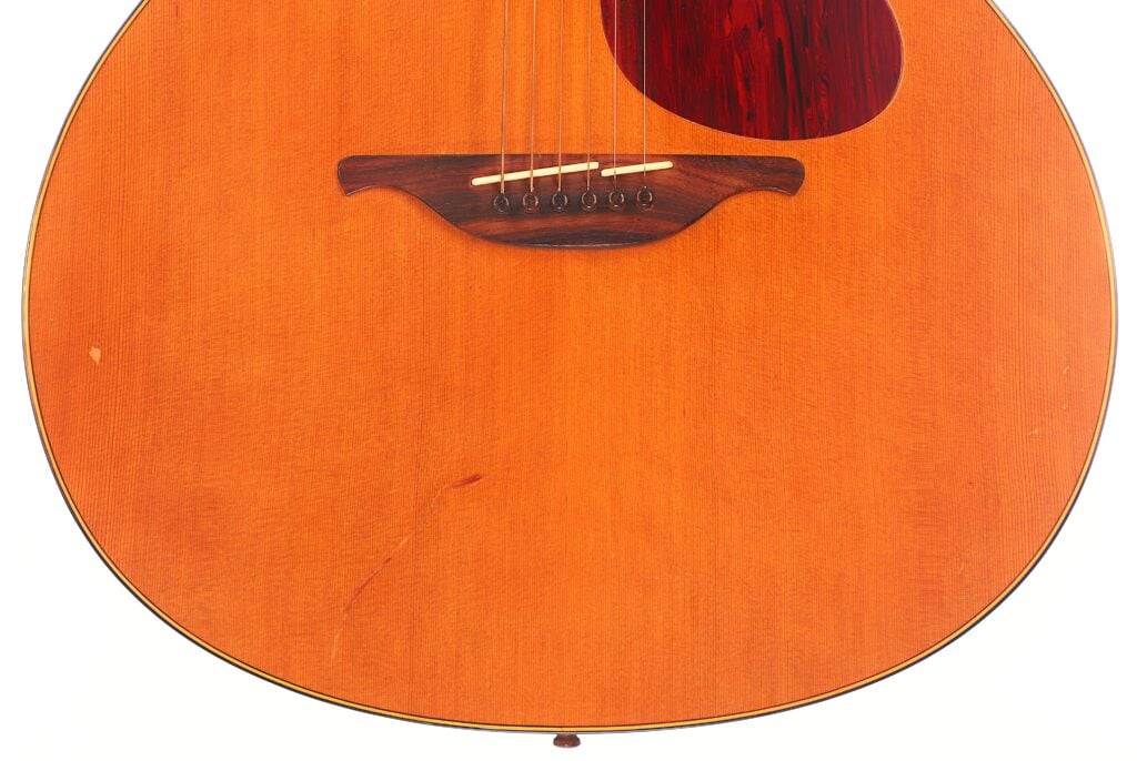 Nahaufnahme des unteren Korpus der Lowden S-5 Akustik Gitarre mit Holzsteg, Saiten und rötlich-braunem Schlagbrett auf einer glatten, orange-braunen Oberfläche. Ein kleiner Kratzer ist in der Nähe der Unterseite sichtbar. Nahaufnahme des unteren Korpus der Lowden S-5 Akustik Gitarre mit Holzsteg, Saiten und rötlich-braunem Schlagbrett auf einer glatten, orange-braunen Oberfläche. Ein kleiner Kratzer ist in der Nähe der Unterseite sichtbar.