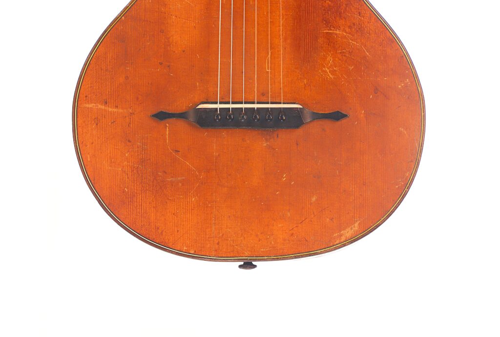 Nahaufnahme der unteren Hälfte einer Braun & Hauser 1905 Klassische Gitarre, die den hölzernen Korpus, die sichtbaren Saiten und den dunklen Steg vor einem weißen Hintergrund zeigt. Nahaufnahme der unteren Hälfte einer Braun & Hauser 1905 Klassische Gitarre, die den hölzernen Korpus, die sichtbaren Saiten und den dunklen Steg vor einem weißen Hintergrund zeigt.