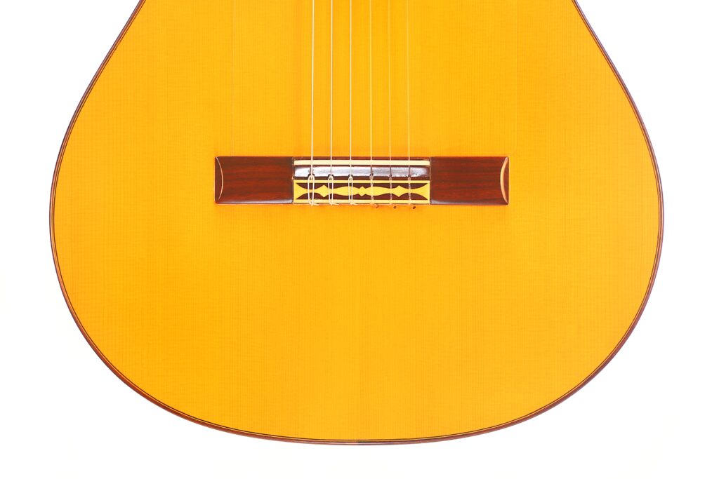Nahaufnahme des Korpus der Juan Montero 1993 Blanca Flamenco Gitarre. Hervorzuheben sind die Saiten, der Holzsteg und die hellbraune Lackierung vor einem weißen Hintergrund. Nahaufnahme des Korpus der Juan Montero 1993 Blanca Flamenco Gitarre. Hervorzuheben sind die Saiten, der Holzsteg und die hellbraune Lackierung vor einem weißen Hintergrund.
