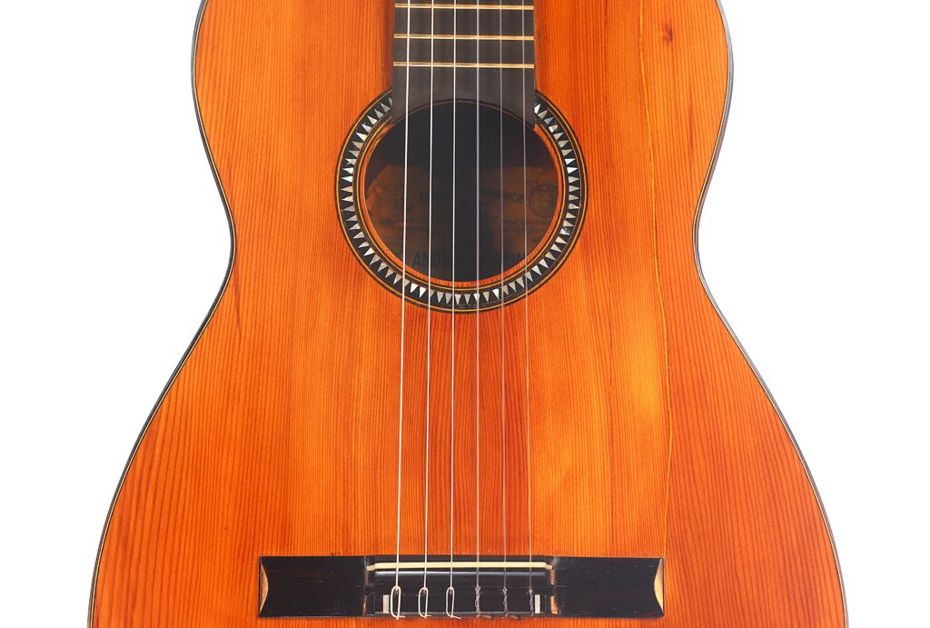 Nahaufnahme des Korpus der Andres Marin 1890 Klassische Gitarre. Hervorzuheben sind die sechs Saiten, die Zierrosette um das Schallloch, der Holzsteg und die polierte Holzoberfläche. Nahaufnahme des Korpus der Andres Marin 1890 Klassische Gitarre. Hervorzuheben sind die sechs Saiten, die Zierrosette um das Schallloch, der Holzsteg und die polierte Holzoberfläche.